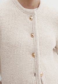 Cremfarbener Strickcardigan mit gerippter Textur, ausgestattet mit fünf goldenen Knöpfen und einem runden Ausschnitt.