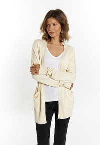 Cardigan lung bej, cu front deschis, textură tricotată, cu două buzunare frontale, purtat peste un top alb cu decolteu în V și pantaloni negri.