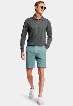 Man loopt en draagt ronde zonnebril, donkergrijs poloshirt met lange mouwen, turquoise korte broek en witte sneakers op een witte achtergrond.