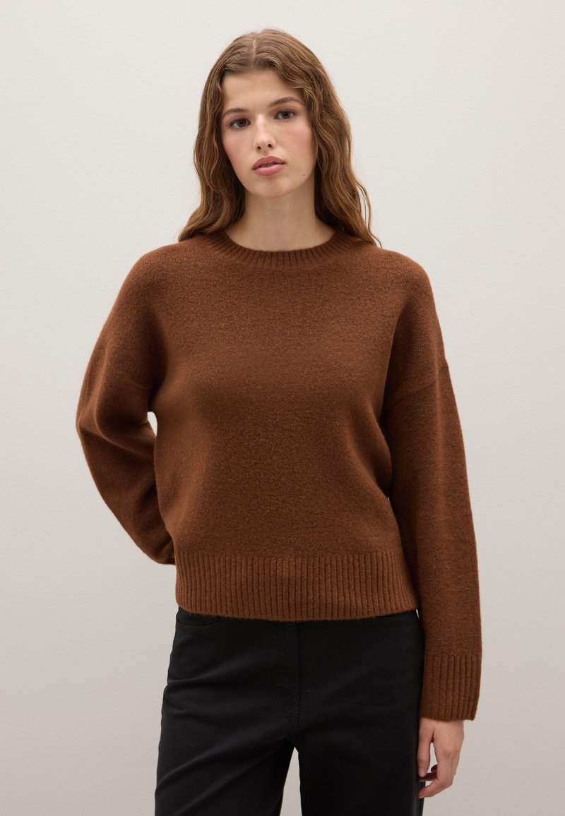 Maglione marrone lavorato a maglia con scollatura rotonda, bordo e polsini a costine. Tessuto morbido e vestibilità rilassata, indossato con pantaloni neri aderenti.