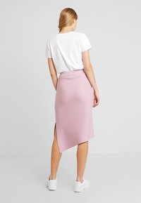 Růžová pletená sukně s asymetrickým lemem a bočním rozparkem, doplněná bílým tričkem a bílými teniskami, zobrazená zezadu.