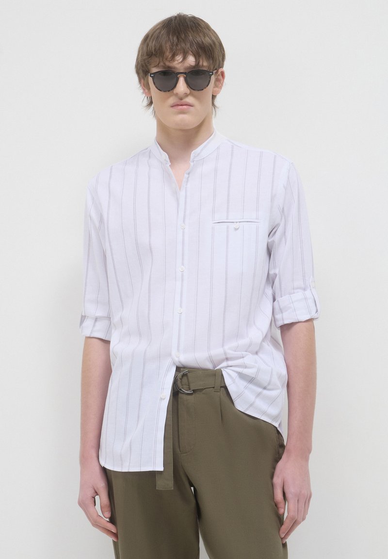 Junger Mann mit schwarzer Sonnenbrille, einem weißen Hemd mit gestreiftem Stehkragen und hochgekrempelten Ärmeln sowie olivgrünen Hosen mit einem Gürtel.