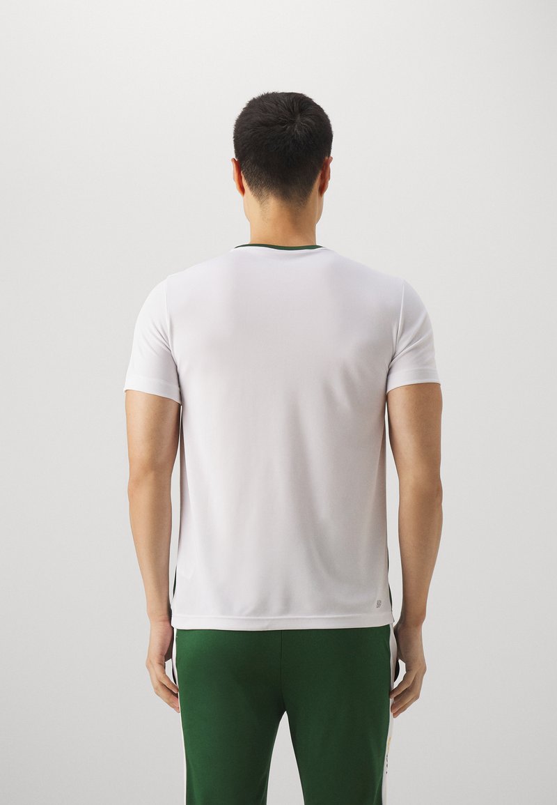 Chemise de sport blanche à manches courtes avec un col vert, design ajusté, texture lisse et accents latéraux. Portée avec un pantalon de sport vert.