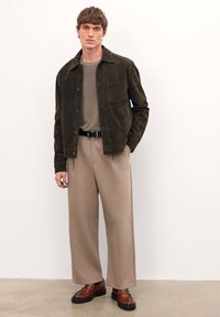 Chaqueta de ante marrón, camisa beis, pantalones anchos beis, zapatos marrones. La chaqueta tiene un bolsillo y detalles de botones, con una textura suave.