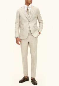 Ljusbeige linnekostym med kavaj med två knappar, matchande byxor, vit skjorta och mönstrad slips. Bruna läderloafers fulländar outfiten.