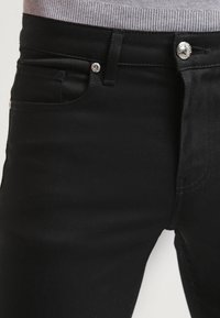 Jeans en denim noir avec un design ajusté, dotés de poches avant, d'une fermeture à bouton en argent et d'une finition lisse et texturée.