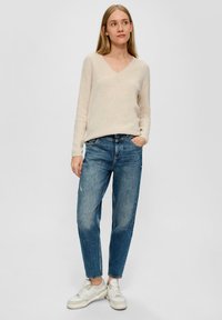 Lichtbeige v-halstrui met geribbelde textuur, gecombineerd met losse blauwe jeans en witte sneakers. Eenvoudig ontwerp met een ontspannen pasvorm.
