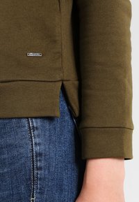 Acercamiento del brazo de una persona que lleva una camiseta de manga larga color verde oliva con un pequeño logo metálico y jeans de mezclilla azul.