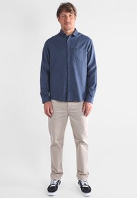 Camisa de manga larga, abotonada, en tejido azul oscuro con un bolsillo. Combinada con pantalones beis claros y zapatillas negras con cordones blancos.