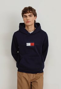 Tamno plava majica s kapuljačom s prednjim džepom; sadrži logotip s crvenim, bijelim i plavim pravokutnicima te tekstom "MY HILFIGER" u sredini.