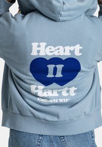 Persona che indossa un felpa azzurro chiaro con la scritta bianca "Heart Hartt" e due cuori blu scuri sovrapposti con "II" sulla schiena.