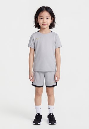 Jeune enfant debout, portant une tenue de sport gris clair avec des logos Nike dorés, des baskets noir et blanc, et des chaussettes blanches Nike sur fond blanc.