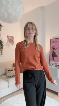 Ein verbranntes Orange, langärmliger Pullover, kombiniert mit hoch taillierten schwarzen Hosen mit einem Knopfdetail. Weiche Texturen und eine lockere Passform.