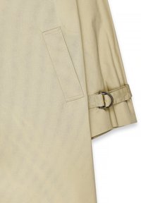 Beige krūšu mētelis ar teksturētu apdari, kas piedāvā sānu kabatu un aproci ar metāla gredzenu. Iekļautas detaļas, piemēram, precīza šūšana.