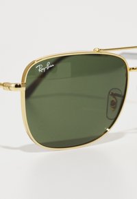 Ochelari de soare din metal aurit cu lentile verzi și formă pătrată. Au temple subțiri și un logo Ray-Ban pe lentilă.