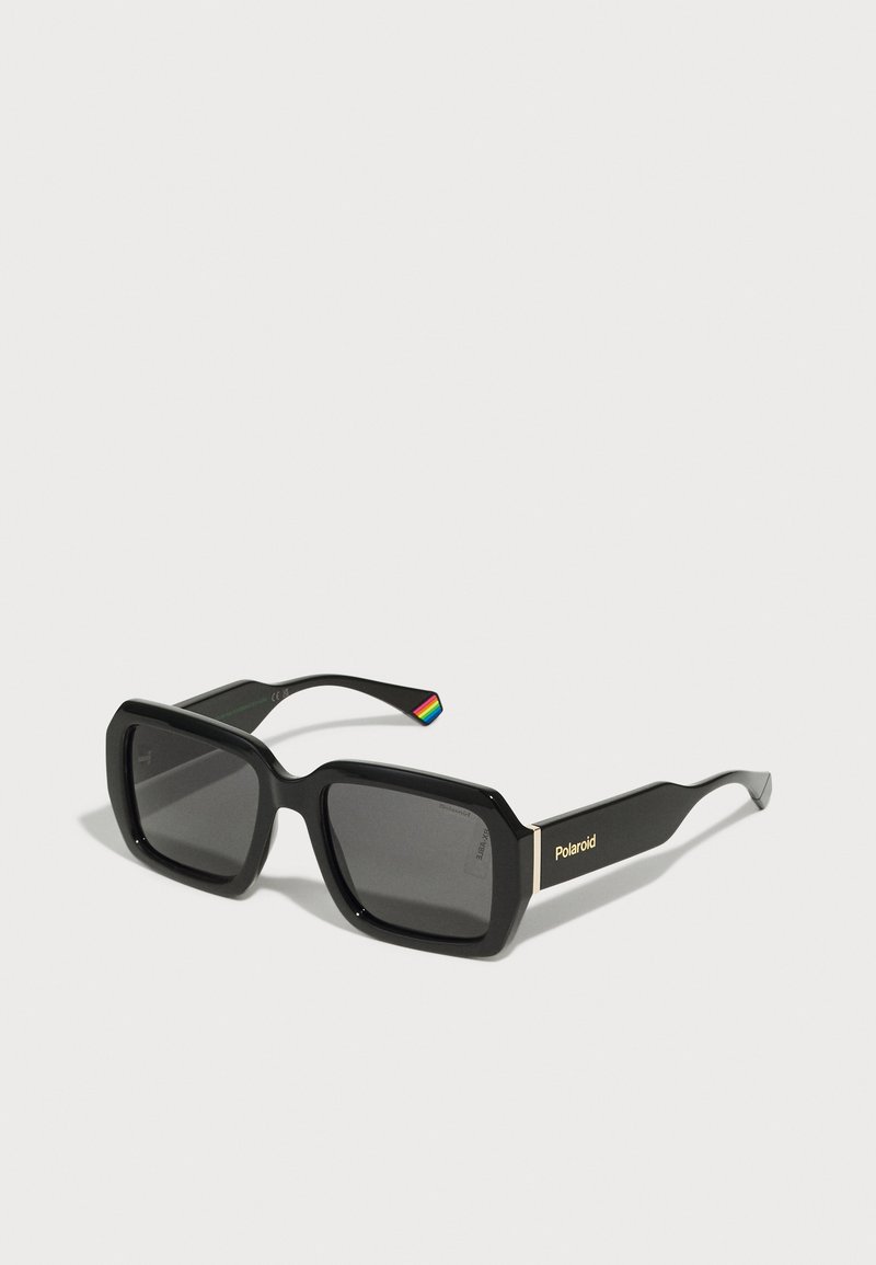 Occhiali da sole neri a forma rettangolare con lenti grigie, montatura in plastica spessa e logo Polaroid dorato sulla tempia. Dettaglio arcobaleno sulla punta dell'asta.