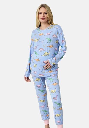 Kvinde iført lyseblå langærmet top og bukser med farverige dinosaurusprint, stående foran en ensfarvet hvid baggrund.