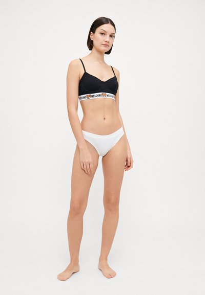 Crna braletica s Moschino logom na elastičnom pojasu, u paru s bijelim bikini gaćicama. Glatka tkanina, minimalistički dizajn. Model pozira protiv jednostavne pozadine.