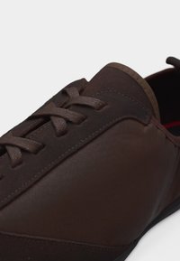 Καφέ δερμάτινο παπούτσι με κορδόνια, στρογγυλεμένη μύτη και λεπτομερή ραφές σε λευκό φόντο, εμφανίζοντας μέρος της σόλας και της γλώσσας στη φτέρνα.