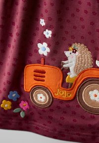 Bordeauxkleurige stof met een geborduurde egeltje op een oranje tractor, omgeven door kleurrijke bloemen. De textuur is zacht met een gestippeld patroon.
