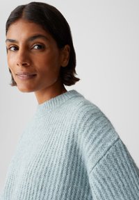 Femme aux cheveux courts et foncés portant un pull en tricot épais bleu clair devant un fond gris uni.