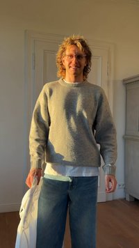 Grauer, strukturierter Pullover mit Rundhalsausschnitt, kombiniert mit locker sitzenden blauen Jeans. Hält eine weiße Tasche und steht vor einer hellen Wand.