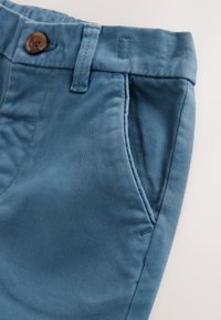 Pantaloni di cotone blu con tasca frontale, passanti per cintura e chiusura con bottone marrone su uno sfondo chiaro.
