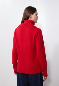Pull en tricot rouge avec un col montant, texture côtelée et coutures verticales à l'arrière. Il présente une coupe décontractée et des manches longues.