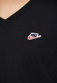 Černé tričko s výstřihem do V vyrobené z hladké bavlny, které má na levé straně hrudi malé vyšití loga Nike v bílé a červené barvě.