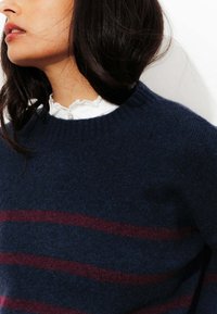 Pull marin bleu avec un col rond, orné de rayures horizontales bordeaux. Fabriqué en tissu doux et texturé. Superposé sur une chemise blanche à col.