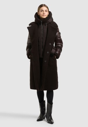 Femme portant un long manteau marron foncé avec une capuche et des manches rembourrées, des leggings noirs et des bottes noires mi-mollet, debout les mains dans les poches.
