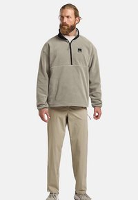 Pull polaire gris clair avec une demi-fermeture éclair et un col noir, associé à un pantalon beige et des baskets de couleur claire. Détail de logo notable.