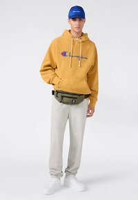 Yellow hoodie with blue text, gray sweatpants, olive green waist bag, and white sneakers; complemented by a blue cap.