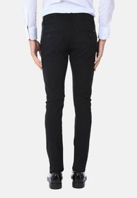 Pantaloni slim-fit neri realizzati in tessuto testurizzato. Presentano due tasche posteriori con chiusura a bottone e passanti per cintura. Indossati con scarpe nere.