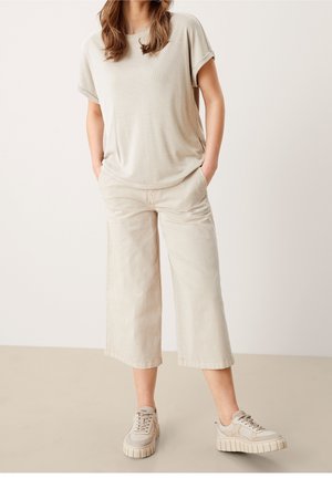 Kobieta ubrana w beżową bluzkę z krótkim rękawem, szerokie spodnie o skróconej długości oraz masywne beżowe trampki, stojąca z rękami w kieszeniach.