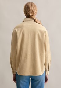 Beżowa koszula sztruksowa z kołnierzykiem, o luźnym kroju, z długimi rękawami i zapinanymi na guziki mankietami, noszona z niebieskimi dżinsami. Widoczny szczegół faktury tkaniny.