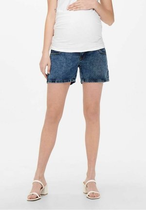 Gravid kvinde iført hvid tanktop, blå graviditetsdenim shorts og hvide hælsandaler, stående på en hvid baggrund.