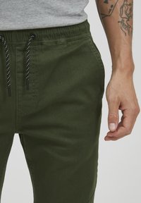 Green cotton joggers with an elastic waistband, adjustable drawstring, and pockets. The fabric has a smooth texture and a matte finish.
