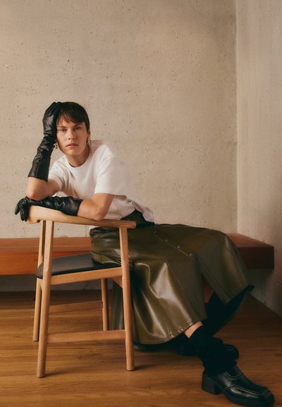 Guanti di pelle nera, t-shirt bianca, gonna di pelle verde con bottoni, seduta su una sedia di legno, contro una parete in cemento testurizzata.