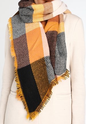 Grande écharpe à carreaux multicolores jaune, noir, marron, gris et orange enroulée autour du cou sur un manteau crème à bords frangés.