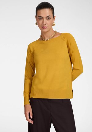 Woman wearing a mustard yellow long-sleeve top and black pants, accessorized with gold hoop earrings and a chunky gold necklace.