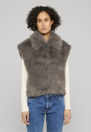 Vrouw met krullend haar, gekleed in een mouwloze grijze faux fur-vest over een crèmekleurig shirt met lange mouwen en een blauwe spijkerbroek, staand tegen een grijze achtergrond.