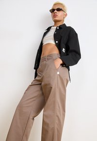 Fekete gombos kabát, fehér crop top, bézs széles nadrág oldalsó zsebekkel. A modellen sötét napszemüveg van, fehér háttér előtt.
