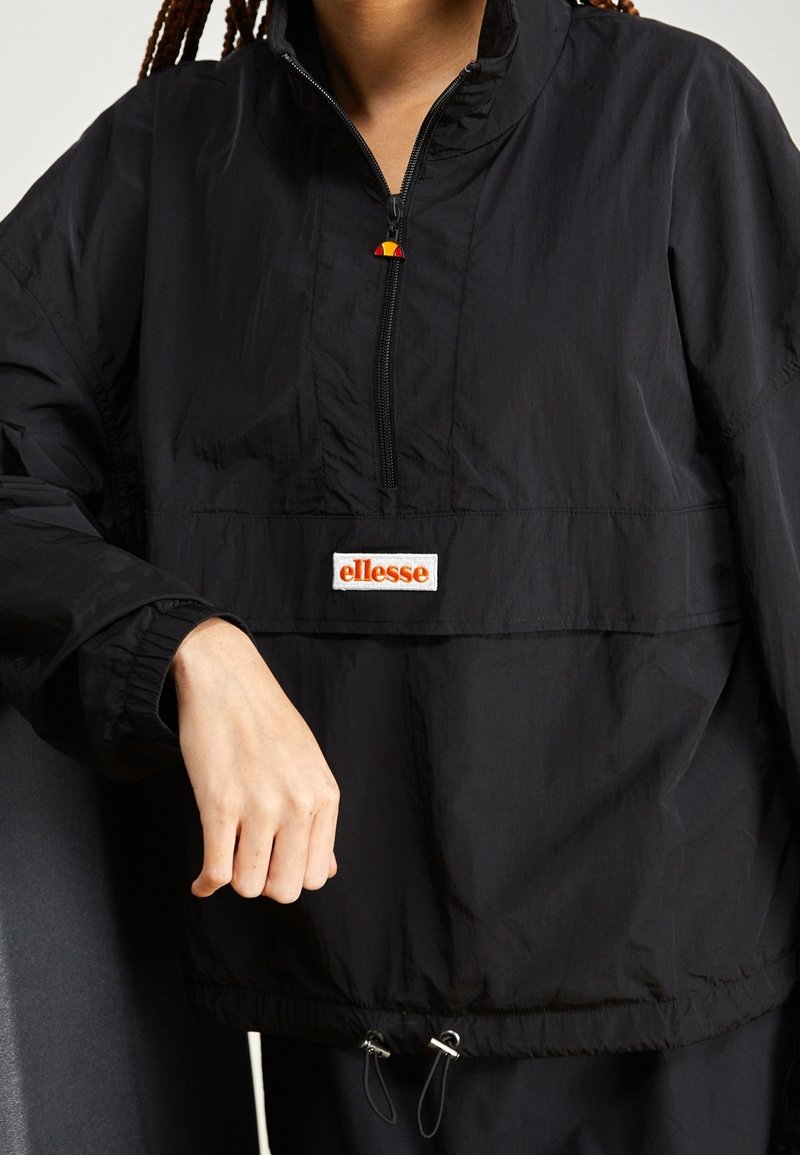 Personne portant une veste à enfiler ellesse noire avec une fermeture éclair à moitié et des cordons, main reposant près de la taille.