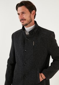Abrigo negro con estampado que presenta un cuello alzado, cierre de botones completo, bolsillos laterales y un bolsillo en el pecho con cremallera, todo con un diseño elegante.