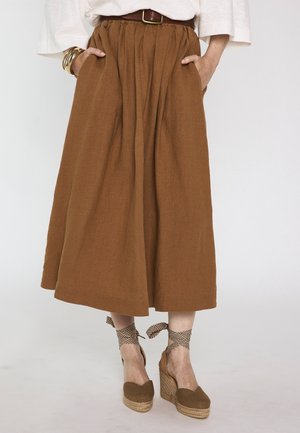 Jupe midi marron avec ceinture, associée à des espadrilles compensées beiges avec liens à nouer autour de la cheville et des bracelets dorés à un poignet.