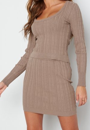 Mujer con un vestido de punto beige acanalado, de manga larga y cuello redondo, mostrado desde el cuello hasta la mitad del muslo sobre un fondo blanco.