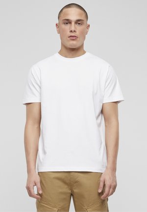 Jeune homme aux cheveux courts portant un t-shirt blanc uni et un pantalon beige, debout devant un fond gris clair.