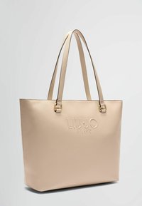 Sac fourre-tout en cuir beige avec double anses, doté d'un logo embossé et d'accents en doré. Texture lisse avec une forme rectangulaire.