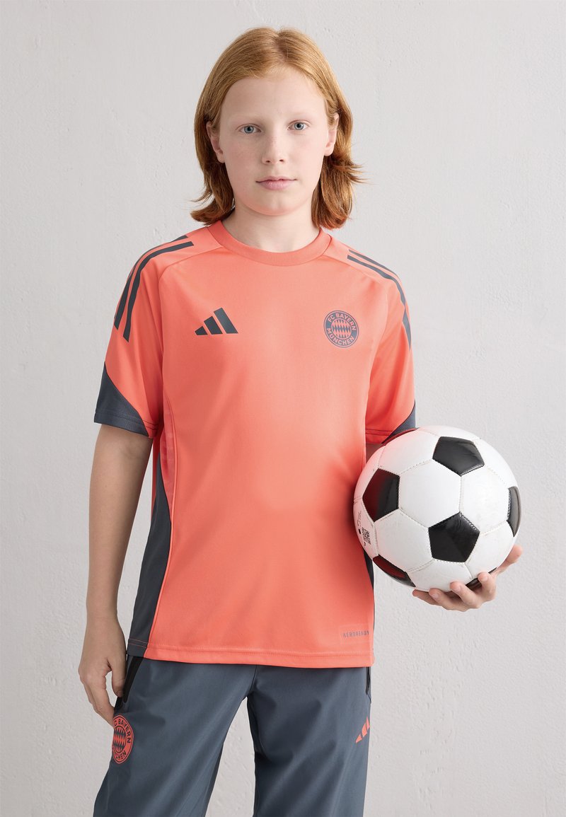 Camisola desportiva da Adidas em laranja brilhante com detalhes em cinza, apresentando o logótipo do FC Bayern Munich. Calças atléticas cinzas e uma bola de futebol preto e branco.